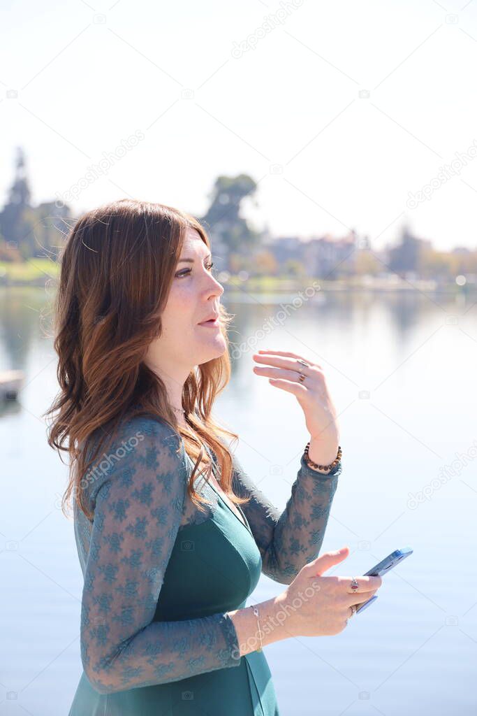 Photo of a Beautiful white girl in a beautiful dress in front of a lake, holding a cell phone