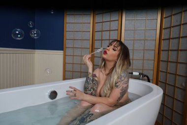 Güzel beyaz bir mankenin küvette oturmuş kabarcıklar üflerken fotoğrafı.