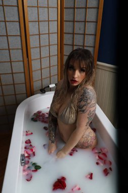 Süt suyu ve gül yapraklarıyla dolu bir küvette oturan güzel beyaz bir mankenin fotoğrafı.