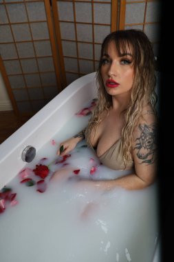 Süt suyu ve gül yapraklarıyla dolu bir küvette oturan güzel beyaz bir mankenin fotoğrafı.