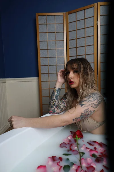 Süt suyu ve gül yapraklarıyla dolu bir küvette oturan güzel beyaz bir mankenin fotoğrafı.