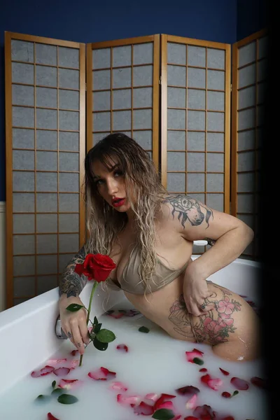 Süt suyu ve gül yapraklarıyla dolu bir küvette oturan güzel beyaz bir mankenin fotoğrafı.