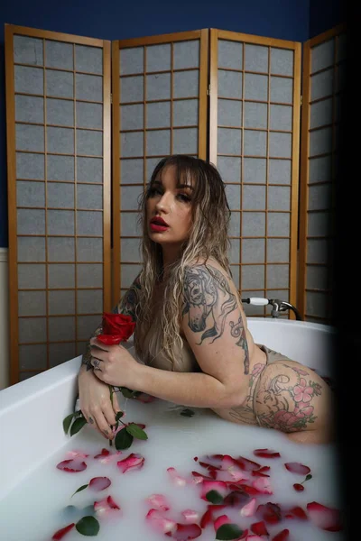 Süt suyu ve gül yapraklarıyla dolu bir küvette oturan güzel beyaz bir mankenin fotoğrafı.