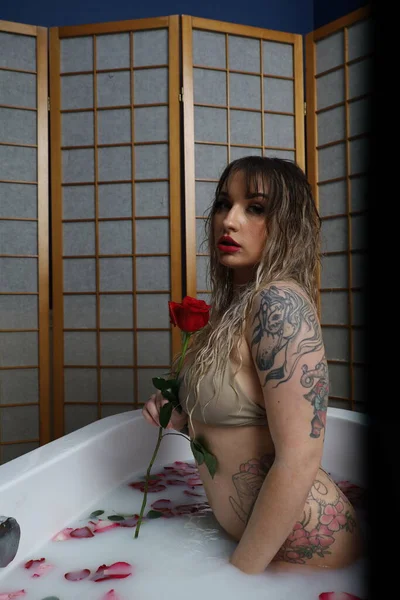 Süt suyu ve gül yapraklarıyla dolu bir küvette oturan güzel beyaz bir mankenin fotoğrafı.