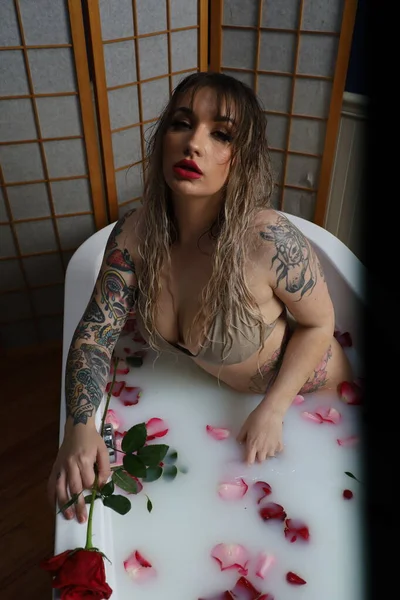 Süt suyu ve gül yapraklarıyla dolu bir küvette oturan güzel beyaz bir mankenin fotoğrafı.