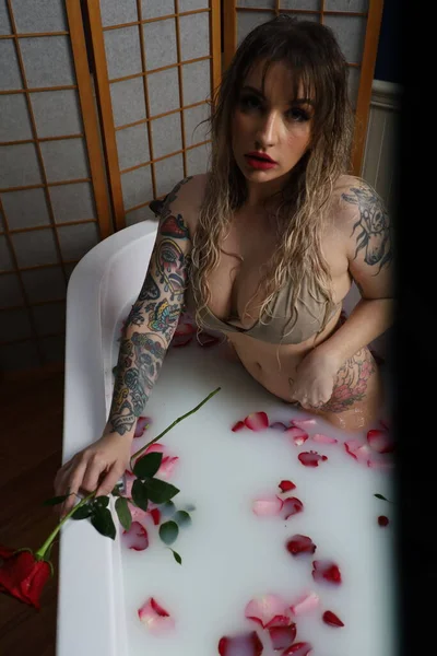 Süt suyu ve gül yapraklarıyla dolu bir küvette oturan güzel beyaz bir mankenin fotoğrafı.