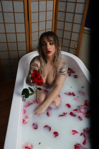 Süt suyu ve gül yapraklarıyla dolu bir küvette oturan güzel beyaz bir mankenin fotoğrafı.