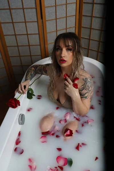 Süt suyu ve gül yapraklarıyla dolu bir küvette oturan güzel beyaz bir mankenin fotoğrafı.