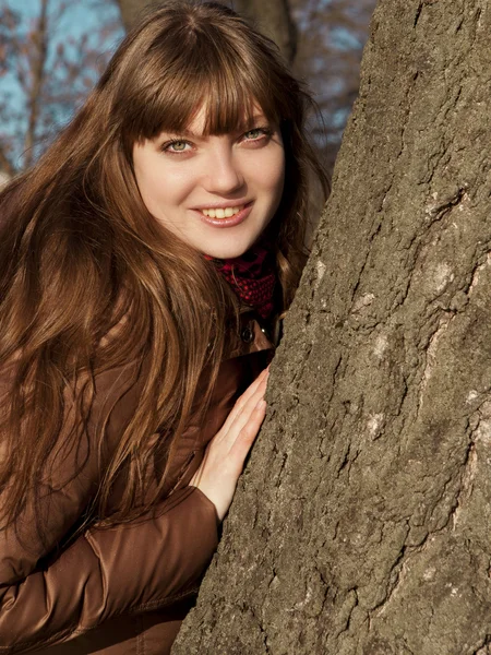 Chica joven con pelo oscuro y abrigo marrón de pie cerca de un árbol — Foto de Stock