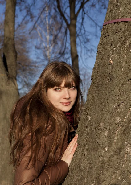 Young girl with dark hair and brown coat standing near a tree — Stock Photo, Image