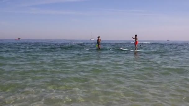 Children ride water sport board SUP on sea coast, boats pass in background — Stock Video