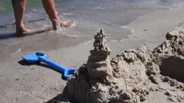 Sand castle and blue toy shovel on beach and barefoot feet of people — ストック動画