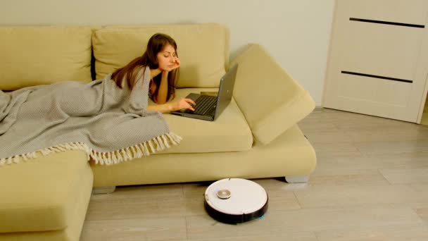Жінка Ноутбуком Лежить Зручному Дивані Час Робот Пилосос Покриває Підлогу — стокове відео