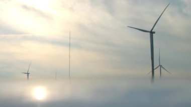 Yel değirmenleri sisli bir günde kıyı istasyonunda elektrik üretir. Büyük pervaneli güçlü makineler gün doğumunda bulutlu gökyüzünün altında döner.