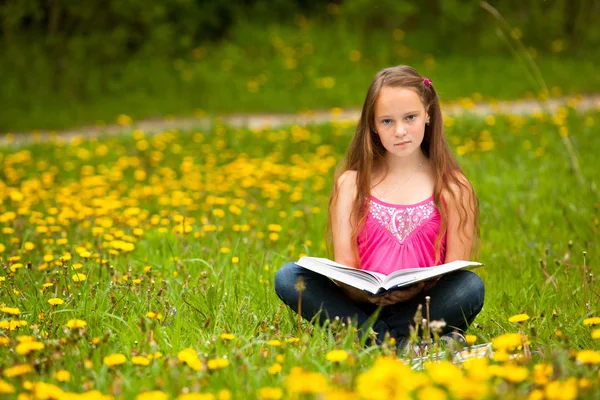 (カメラを見る) の女の子は、草原の本を読みます — ストック写真