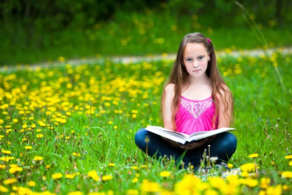 Teenie-Mädchen (blickt in die Kamera) liest ein Buch auf der Wiese Stockbild