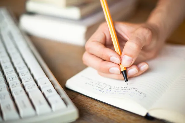 Photo of hands writes a pen in a notebook, computer keyboard and a stack of books in background Stock Picture