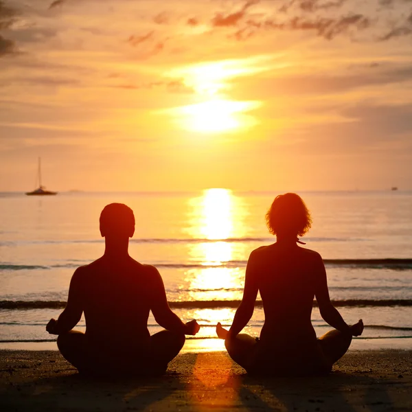 Практика йоги, молодая пара, сидящая на берегу моря в позе лотоса на закате — стоковое фото