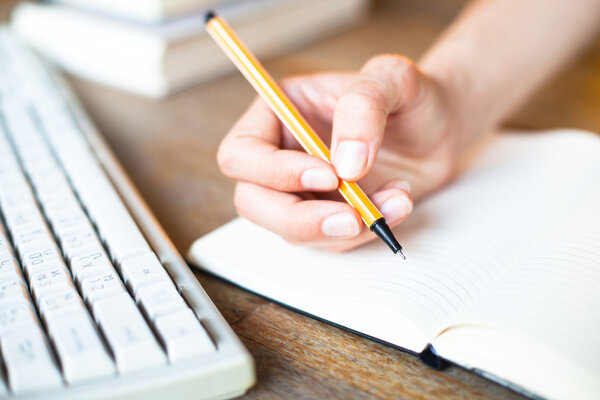 Hands writes a pen in a notebook (computer keyboard, a stack of books in background)