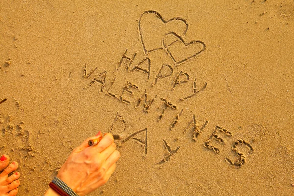 Female hand writes in the beach sand: Happy Valentine's Day and draws two hearts — Stock Photo, Image