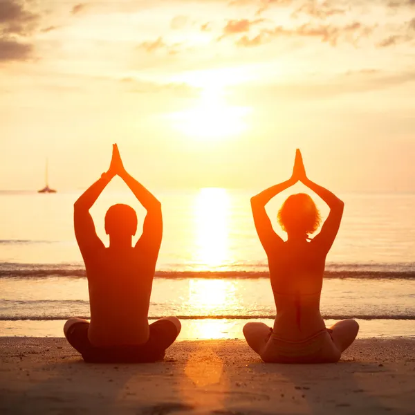 Prática de ioga, jovem casal sentado na praia do mar na posição de lótus ao pôr do sol . — Fotografia de Stock