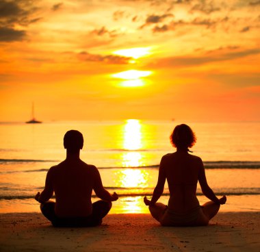 A young couple sitting on the beach of the sea in the lotus position at sunset, yoga practice.