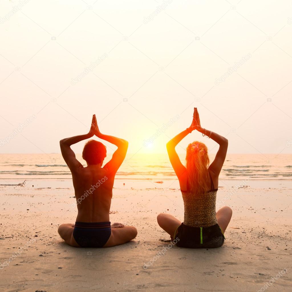Young couple sitting on the beach of the sea in the lotus position at sunset