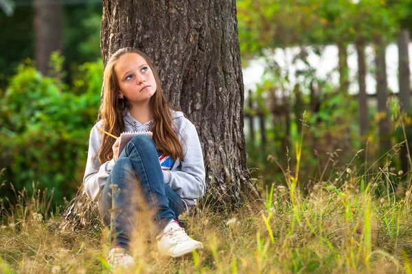 Dospívající dívka psaní v poznámkovém bloku, zatímco sedí v parku. Stock Fotografie