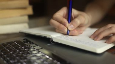 genç bir kadının yakın çekim bir kalem bir dizüstü bilgisayar, dizüstü bilgisayar ve arka planda kitaplar yazıyor