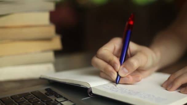 Close-up van een jonge vrouw schrijft een pen in een notitieboekje, laptop en boeken in de achtergrond — Stockvideo