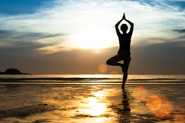 Silhuetten av en ung kvinna som utövar yoga på stranden vid solnedgången. — Stockfoto