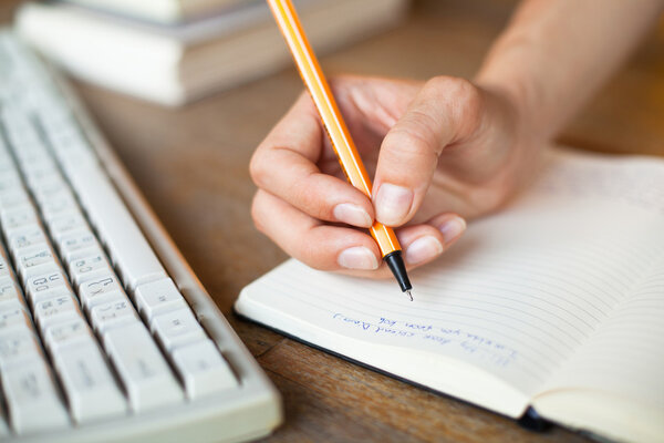 Hands writes a pen in a notebook, computer keyboard in background