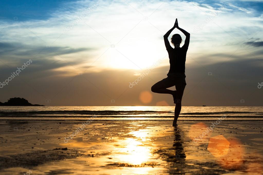 Silhouette of a young woman practicing yoga on the beach at sunset.