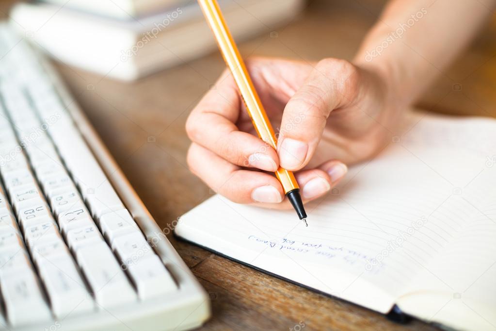 Hands writes a pen in a notebook, computer keyboard in background.