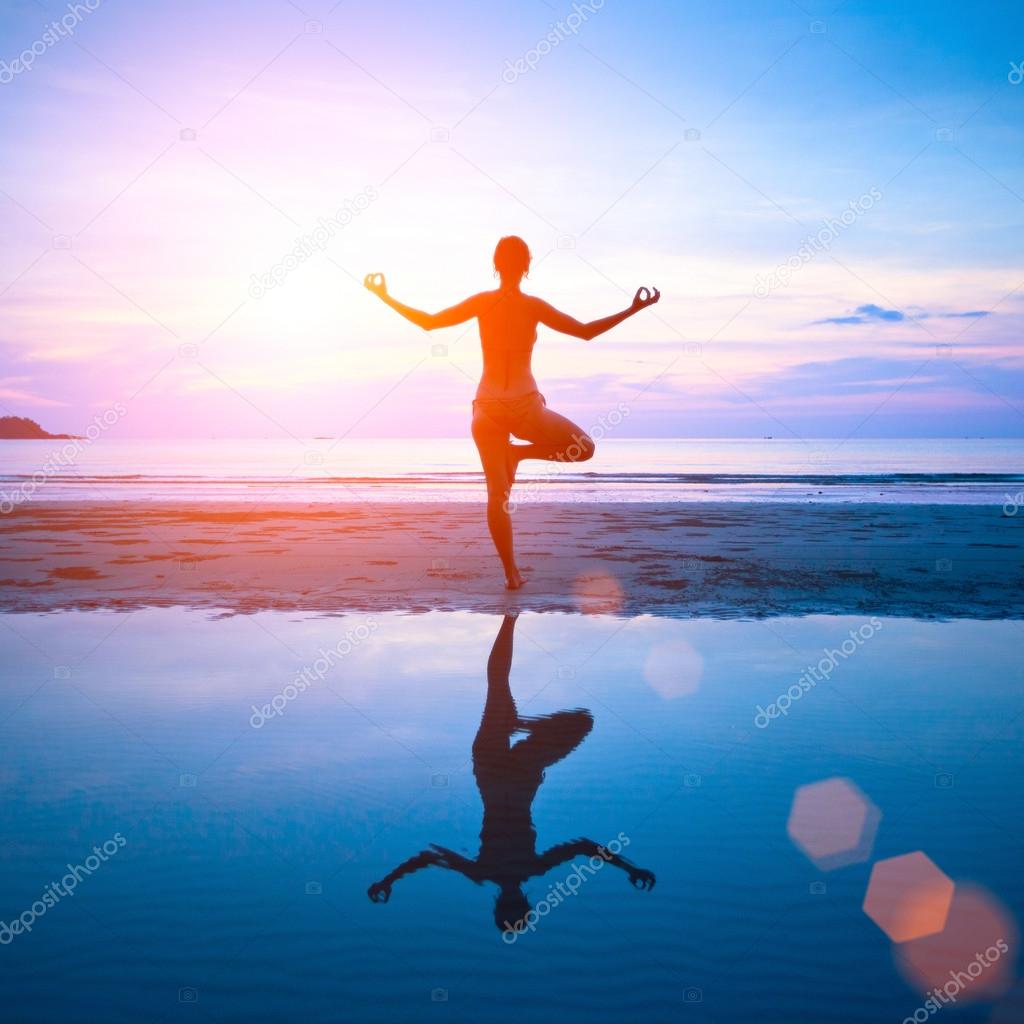 Young woman practicing yoga on the beach at sunset with reflection in water.