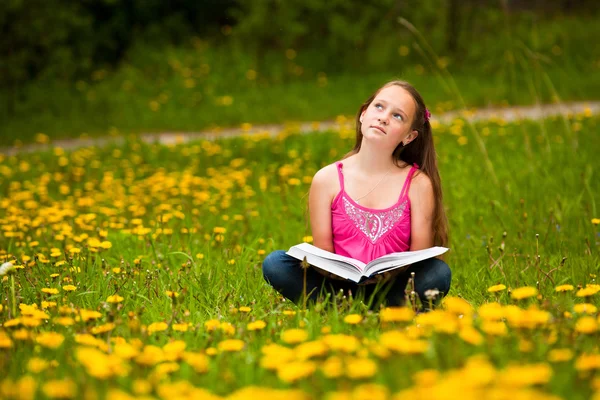 Menina senta-se em uma grama e sonha enquanto lê um livro — Fotografia de Stock