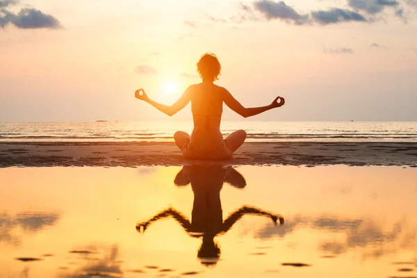 Yoga vrouw in lotus pose zittend op het strand tijdens zonsondergang, met weerspiegeling in water. — Stockfoto