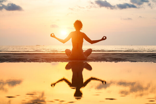 Yoga woman sitting in lotus pose on the beach during sunset, with reflection in water.