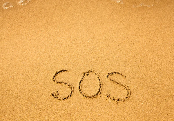 SOS - scritto in sabbia sulla texture della spiaggia, morbida onda del mare . — Foto Stock