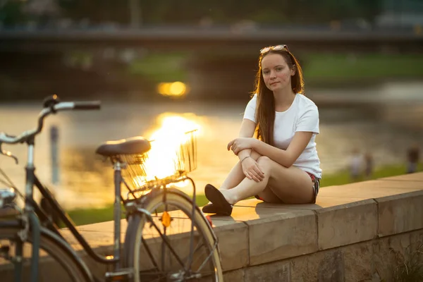 かわいい女の子と自転車は素晴らしい夕日で川の遊歩道に座っている — ストック写真