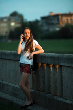 Genç bir kız telefonda konuşuyor şehrin nehir kıyısında duruyor..