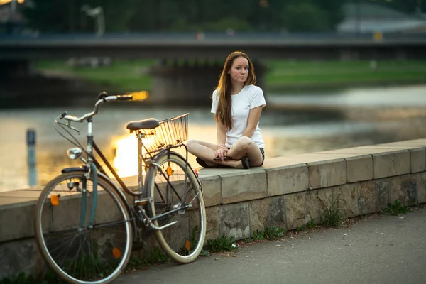 街の川のほとりに自転車を持つ10代の少女が座っている — ストック写真