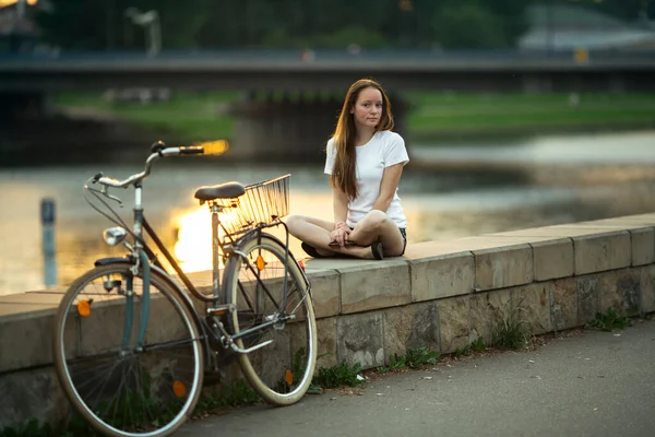 街の川のほとりに自転車を持つ10代の少女が座っている ロイヤリティフリーのストック写真