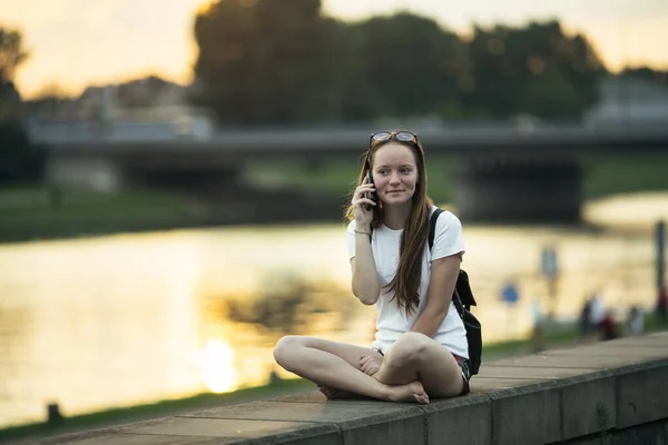 日没時に街のウォーターフロントに座って電話で話している女の子 — ストック写真