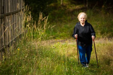 Yaşlı bir kadın sopalarla yürüyor, kırsalda yürüme alıştırması yapıyor..
