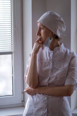 Yüzünde maske olan bir kadın doktor endişeyle pencereden dışarı bakar..