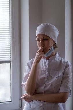 Yüzünde maske olan kadın bir doktor pencerenin yanında durup endişeyle bakıyor..