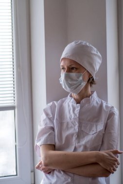 Pencere kenarında duran maske takan kadın bir doktor..