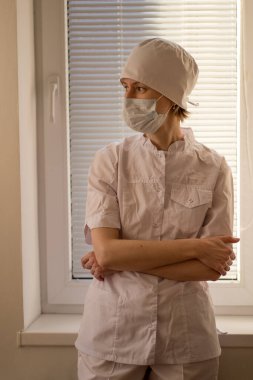 Yüz maskesi takan bir doktor endişeyle hastane penceresinin yanında duruyor..