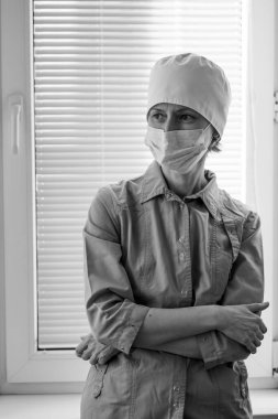 Pencerenin yanında duran yüz maskeli bir kadın doktor. Siyah beyaz fotoğraf.